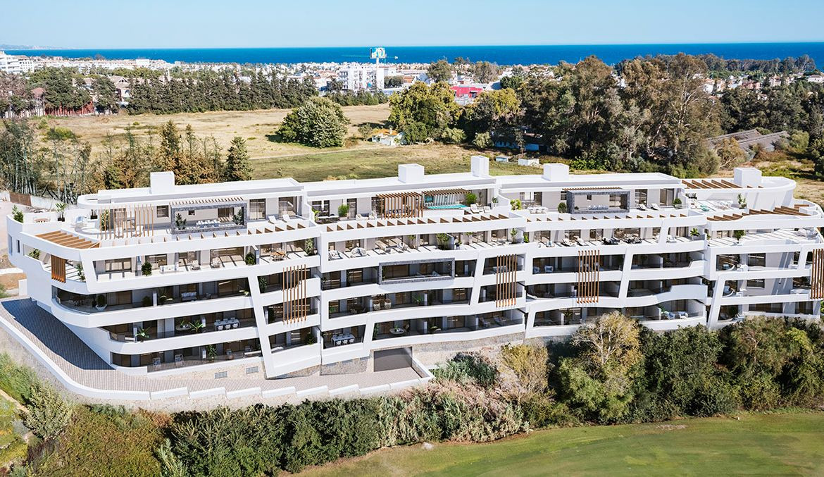 Modern white apartment building with terraces, overlooking lush greenery and the sea in a coastal location.